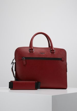 Mallette en cuir texturé rouge avec poignées supérieures, poche avant zippée et bandoulière amovible, exposée sur un piédestal blanc.