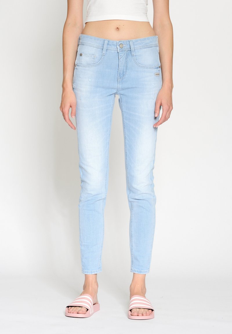 Light blue skinny jeans made of denim with a faded finish, featuring front pockets, a zip fly, and a fitted silhouette. Paired with pink slide sandals.
