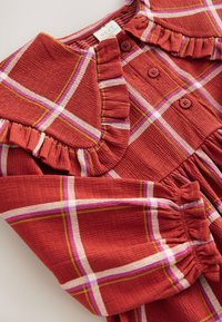 Vestito a quadri rossi con colletto arricciato, dotato di bottoni e maniche arricciate. Il tessuto ha una finitura strutturata con accenti bianchi e rosa.