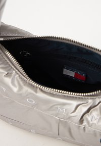 Metāliski sudraba pleca soma, izgatavota no gludas materiāla, ar rāvējslēdzēju, melnu oderi un mazu etiķeti ar uzrakstu "TOMMY JEANS."