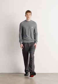 Pulover gri din tricot cu guler rotund și un logo mic, asortat cu pantaloni de pijama în dungi închise. Modelul poartă pantofi negri cu talpă roșie.