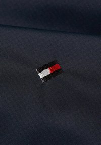 Tessuto blu navy con un piccolo logo bandiera ricamato in rosso, bianco e nero. Texture liscia con un leggero scintillio.