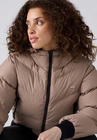 Chaqueta de plumas en taupe claro con acolchado en forma de chevrón, cierre frontal y puños de canalé negros. Cuenta con un alto cuello y un discreto detalle de logo.