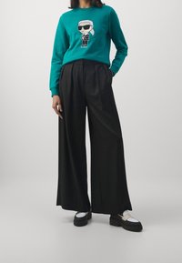 Sudadera azul verdosa con gráfico de dibujos animados en el centro, combinada con pantalones de pierna ancha negros. El calzado presenta una suela gruesa con un acento blanco.