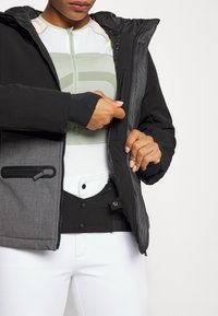 Osoba w warstwowym stroju sportowym i kurtce w kolorowe bloki, dostosowująca zamek błyskawiczny wewnątrz kurtki obiema rękami.