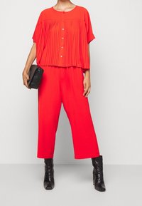 Top rojo plisado con escote redondeado y cierre de botones, combinado con pantalones rojos holgados y de pierna ancha, y botas negras hasta el tobillo.