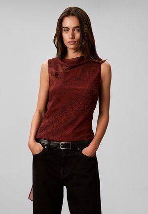 Femme aux longs cheveux bruns portant un haut sans manches rouge foncé à motifs et un pantalon noir, les mains dans les poches, sur un fond gris uni.
