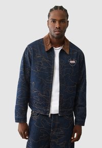 Spijkerjack met een bruine corduroy kraag, blauwe stof met een oranje abstract patroon en een ritssluiting aan de voorkant.