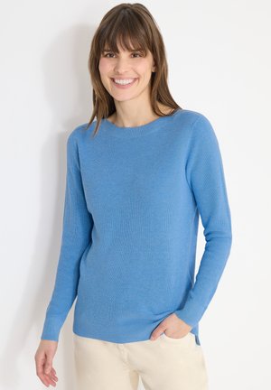 Donna sorridente con capelli castani che indossa un maglione di maglia azzurro chiaro e pantaloni color crema, in piedi davanti a uno sfondo bianco.