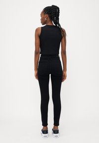 Fekete cropped tank top, magas derekú fekete skinny farmerrel és fekete-fehér sportcipővel párosítva; a modell hátulról látható.