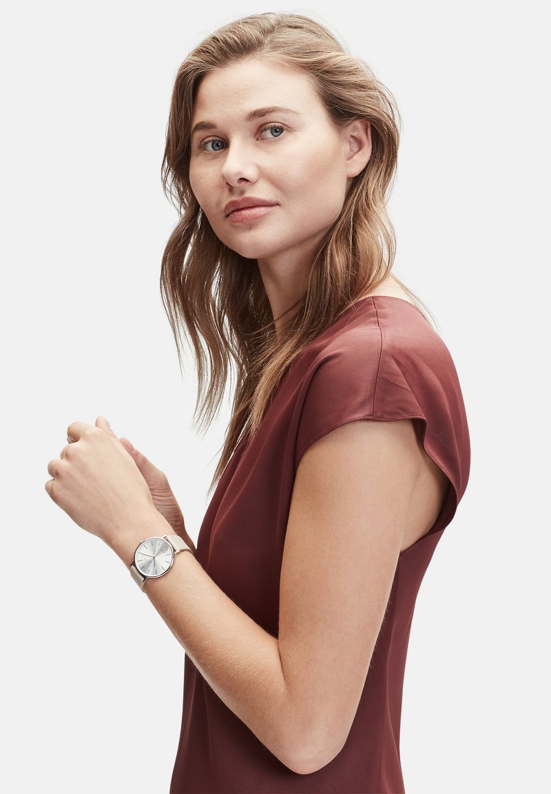 Zilveren polshorloge met een ronde wijzerplaat en minimalistisch ontwerp, gedragen om een pols. De drager heeft lang, golvend haar en is gekleed in een kastanjebruine top.