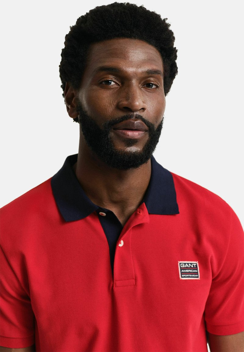 Rotes Polohemd aus Baumwolle mit navyfarbenem Kragen, versehen mit einem kleinen GANT-Logo-Patch und zwei Knöpfen an der Knopfleiste. Schlichtes Design.