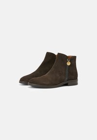 Bottines en daim brun foncé avec des petits talons, garniture latérale tressée et une petite étiquette ronde en or sur fond blanc uni.