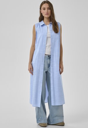 Kobieta w niebieskiej, bezrękawnej, długiej kamizelce w paski, zapinanej na guziki, nosząca biały top i luźne, szerokie, jasnoniebieskie dżinsy, stojąca na szarym tle.