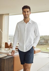 Camicia bianca in lino con maniche lunghe e chiusura a bottoni, abbinata a pantaloni corti blu scuro. Design moderno e minimalista in un ambiente luminoso e interno.