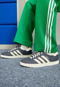 Grå canvas sneakers med vita ränder och gummisålar, kombinerat med gröna byxor med vita sidostripes, på en heltäckningsmatta.