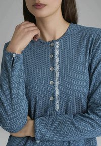 Top a maniche lunghe con motivo blu, dotato di una chiusura anteriore con bottoni, rifiniture in pizzo lungo la pettorina e polsini fitted con chiusure a bottone.