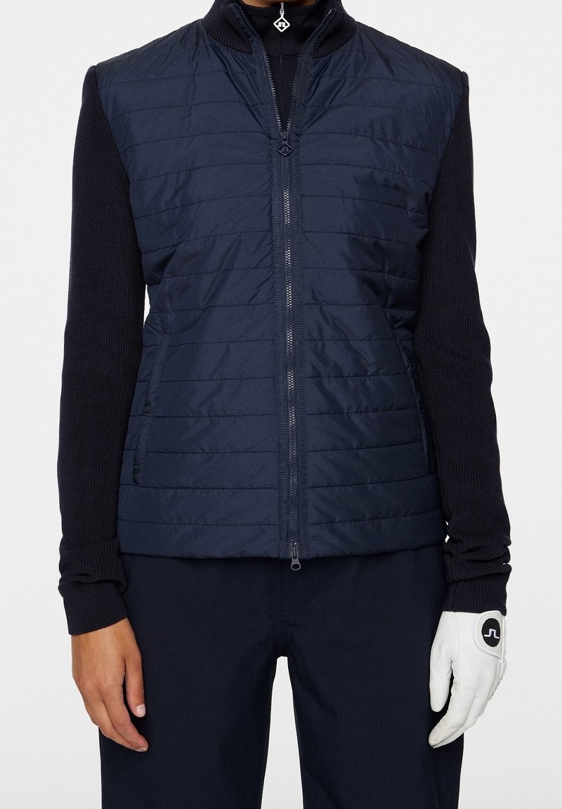 Marinblå puffväst med dragkedja och mönstrad textur, kombinerad med en figurnära svart långärmad tröja och vita handskar med logotyp.