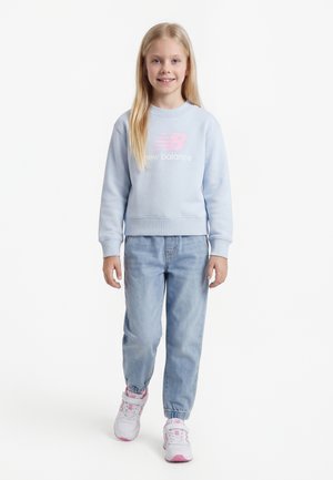 Chica con cabello largo y rubio, vistiendo una sudadera azul claro de New Balance, jeans azul claro y zapatillas blancas con rosa, de pie y sonriendo.