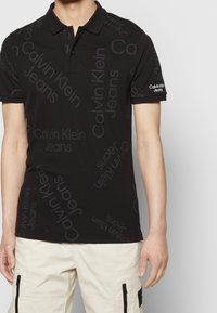 Ung person som bär en svart Calvin Klein Jeans-piké med upprepande logotext och ljusa cargobyxor med dragkedjefickor.