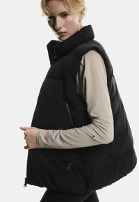 Sort puffy vest med høj krave og lynlåslukning, som har en stor frontlomme, glat tekstur og fremtrædende branding på siden.