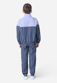 Modra trenirka z svetlo modrim zgornjim delom in temno modrim dnom. Ima geometrijski beli vzorec, elastični pas in manšete. Enobarvni beli čevlji.