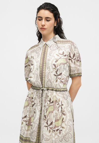 Mujer con un vestido crema estampado con diseños de pájaros y hojas, mangas cortas, cuello y cintura ajustable con cordón, mirando hacia abajo.