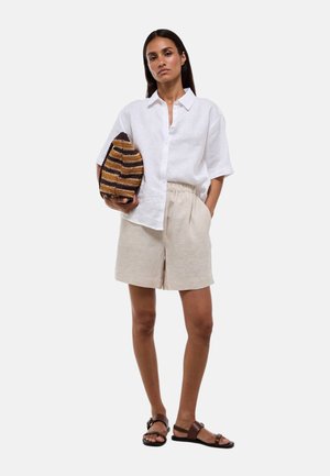 Femme portant une chemise blanche à manches courtes boutonnée, un short beige, des sandales marron, tenant une pochette rayée marron et jaune avec une main dans la poche.