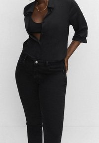 Camisa negra de botones con mangas enrolladas, superpuesta a una camiseta de tirantes negra. Combinada con jeans negros ajustados, mostrando un look monocromático y elegante.