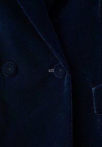 Blazer de terciopelo azul marino oscuro de doble botonadura con dos botones redondos y detalles de costura visibles en las solapas.