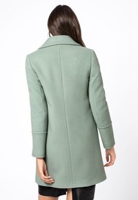 Zelený kabát ze směsi vlny s přiléhavým střihem, dlouhými rukávy a klopovým límcem. Vyznačuje se rovnými zády a délkou do poloviny stehen.