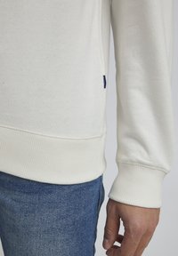 Sudadera de color crema claro con puños y dobladillo acanalados, que presenta una textura sutil. Combinada con jeans de mezclilla, mostrando un ajuste casual.