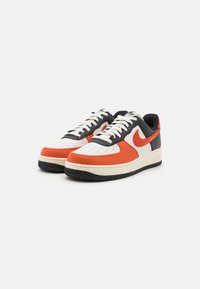 Las zapatillas Nike Air Force 1 presentan una parte superior de cuero blanco con acentos en naranja y marino, suela de goma negra y un diseño clásico de puntera perforada.