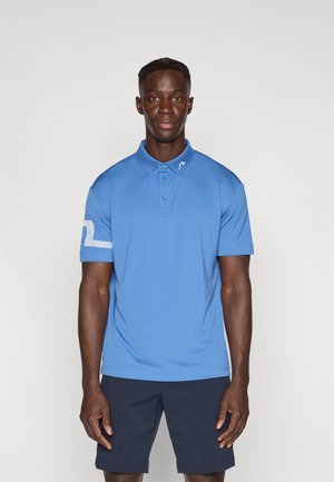 Modré polo tričko z hladkej látky s golierom, zapínaním na tri gombíky a prúžkovaným detailom na rukáve. Nosí sa s námorníckymi šortkami.