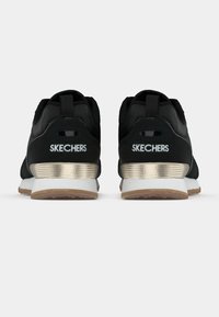 Par de sapatilhas pretas Skechers vistas de trás, com entressolas brancas e detalhes em dourado metálico no calcanhar, em fundo branco.