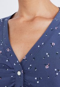 Gros plan sur un chemisier à col en V bleu à motifs floraux, avec un seul bouton blanc et de légères fronces au centre devant.