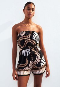 Une combinaison noire sans bretelles mettant en avant de grands motifs floraux et géométriques en beige et blanc, avec une taille cintrée et un détail de ceinture nouée.