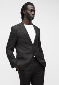 Άνδρας με κοτσίδες φορώντας σκούρο καφέ κοστούμι και λευκό μπλουζάκι, στέκεται με τα χέρια στις τσέπες, κοιτώντας προς τα πλάγια μπροστά σε απλό φόντο.