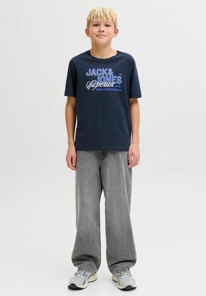 Temno modra bombažna majica z belim in modrim grafičnim napisom spredaj, kombinirana z ohlapnimi sivimi jeans hlačami in sivimi supergami.