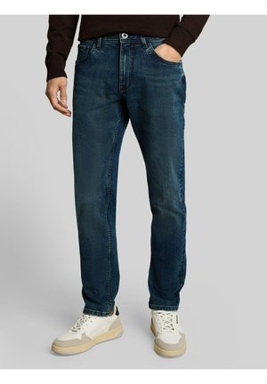Man draagt donkerblauwe rechte jeans met voorzakken, gecombineerd met witte en beige sneakers tegen een effen grijze achtergrond.