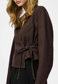 Brun slå-om bluse med lange ærmer og bindebånd i taljen, med dyb V-hals og glat tekstur. Brugt med sort påklædning.