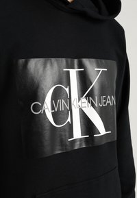 Felpa con cappuccio nera, caratterizzata da una grafica lucida e sovradimensionata con il testo "CK" e "CALVIN KLEIN JEAN" in bianco. Il tessuto sembra liscio.