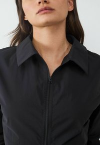 Schwarze Zip-Up-Jacke mit großem Kragen, leichtem Stoff mit leicht texturierter Oberfläche und zentralem Reißverschluss.