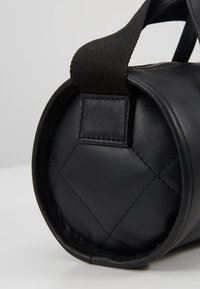 Extrémité ronde d'un sac en cuir noir matelassé avec une bandoulière tissée noire fixée par une couture rectangulaire.