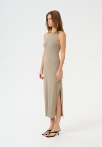 Beiges, ärmelloses Kleid mit tailliertem Design, seitlichen Schlitzen und glatter Textur. Kombiniert mit schwarzen Flip-Flop-Sandalen.