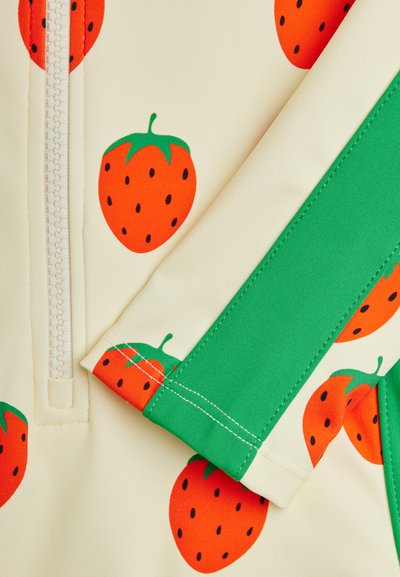 Κοντινό πλάνο κρεμ υφάσματος με κόκκινα σχέδια φράουλας, πράσινες λεπτομέρειες φύλλων, αντιθετική πράσινη ρίγα, λευκό φερμουάρ και ορατές λεπτομέρειες ραφής.