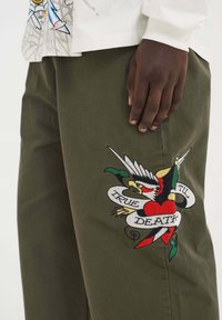Groene katoenen broek met een kleurrijk geborduurd ontwerp van een vogel, een hart en een banner met de tekst "TRUE TIL DEATH."