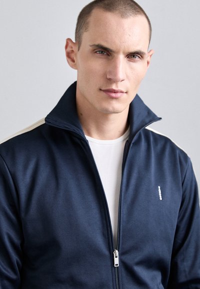 Ung man med kort hår som bär en marinblå zip-up jacka över en vit skjorta mot en enkel grå bakgrund.