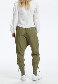 Mujer con una blusa smock blanca de manga larga, pantalones holgados en verde oliva y zapatillas blancas, de pie contra un fondo liso.