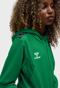 Grøn hættetrøje med lynlås lavet af tekstureret stof, med et sort skulderdesign og en hvid logo på brystet.